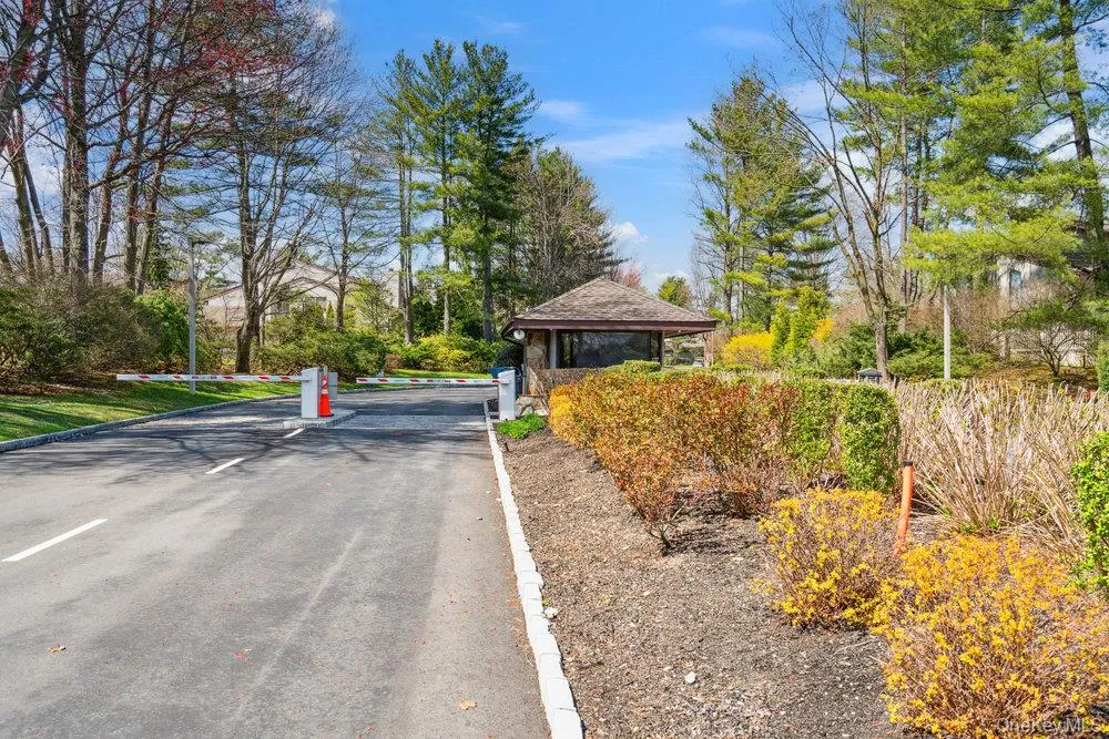 Gated entry featuring an attendant booth with a distinct shingled roof, asphalt driveway, automated entry arm, and surrounding mature landscaping Gated entry featuring an attendant booth with a distinct shingled roof, asphalt driveway, automated entry arm, and surrounding mature landscaping