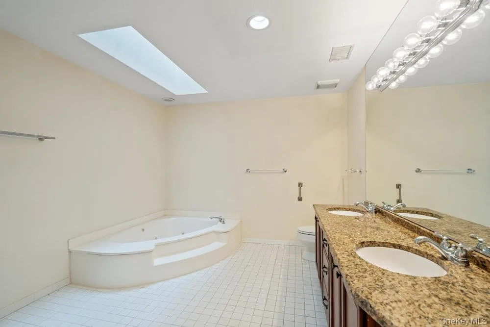 Bathroom featuring a corner jetted tub with steps, a rectangular skylight, and a double vanity with a granite countertop and dual undermount sinks Bathroom featuring a corner jetted tub with steps, a rectangular skylight, and a double vanity with a granite countertop and dual undermount sinks