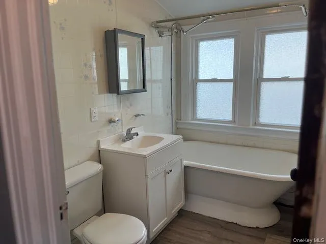 Bathroom featuring wood-type flooring, tile walls, a tub to relax in, vanity, and toilet Bathroom featuring wood-type flooring, tile walls, a tub to relax in, vanity, and toilet