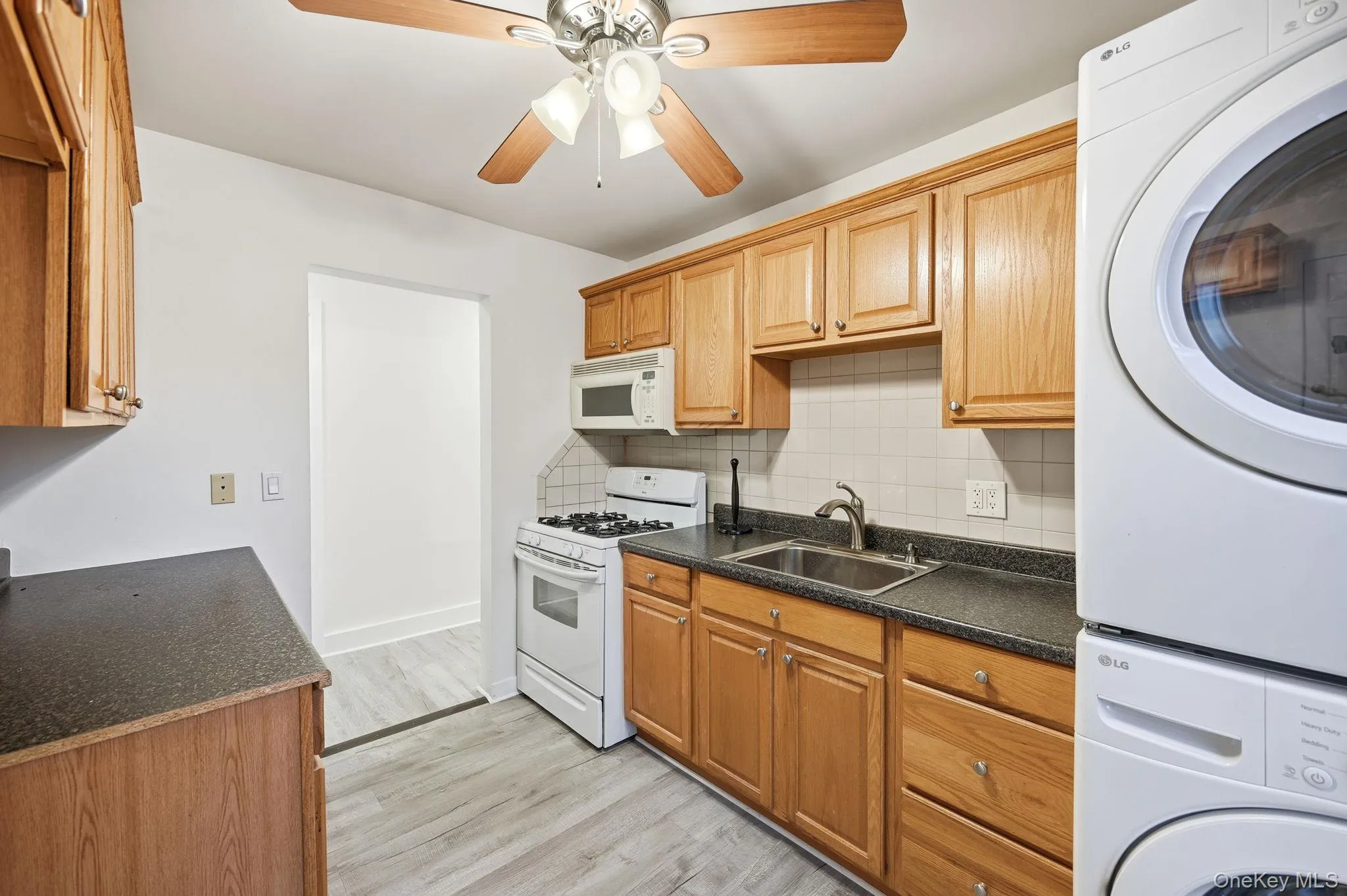 Kitchen with white appliances, dark countertops, ceiling fan, decorative backsplash, and light wood finished floors Kitchen with white appliances, dark countertops, ceiling fan, decorative backsplash, and light wood finished floors