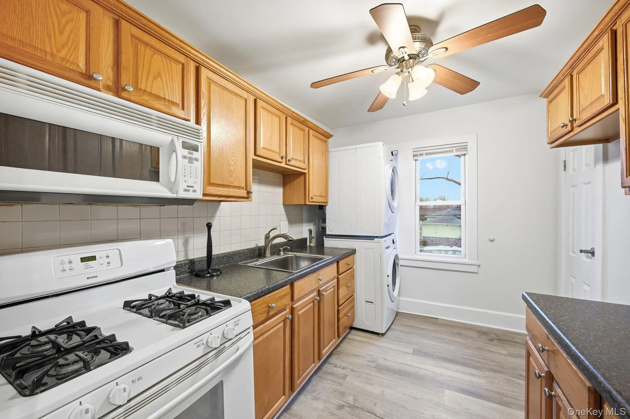Kitchen with white appliances, stacked washing machine and dryer, tasteful backsplash, light wood finished floors, and ceiling fan Kitchen with white appliances, stacked washing machine and dryer, tasteful backsplash, light wood finished floors, and ceiling fan