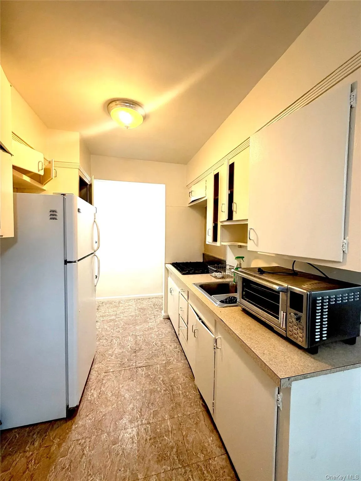 Galley kitchen featuring a white refrigerator, white cabinetry, a built-in sink, a stovetop, and patterned flooring Galley kitchen featuring a white refrigerator, white cabinetry, a built-in sink, a stovetop, and patterned flooring