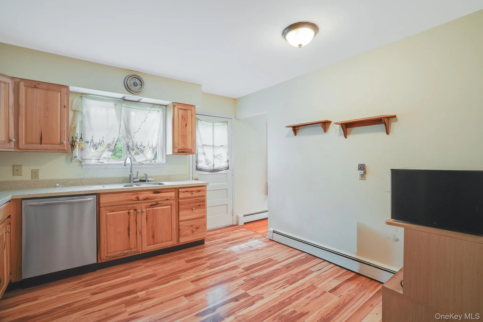 Kitchen with wood-finish flooring, natural wood cabinetry, light-toned countertops, and a stainless steel dishwasher Kitchen with wood-finish flooring, natural wood cabinetry, light-toned countertops, and a stainless steel dishwasher