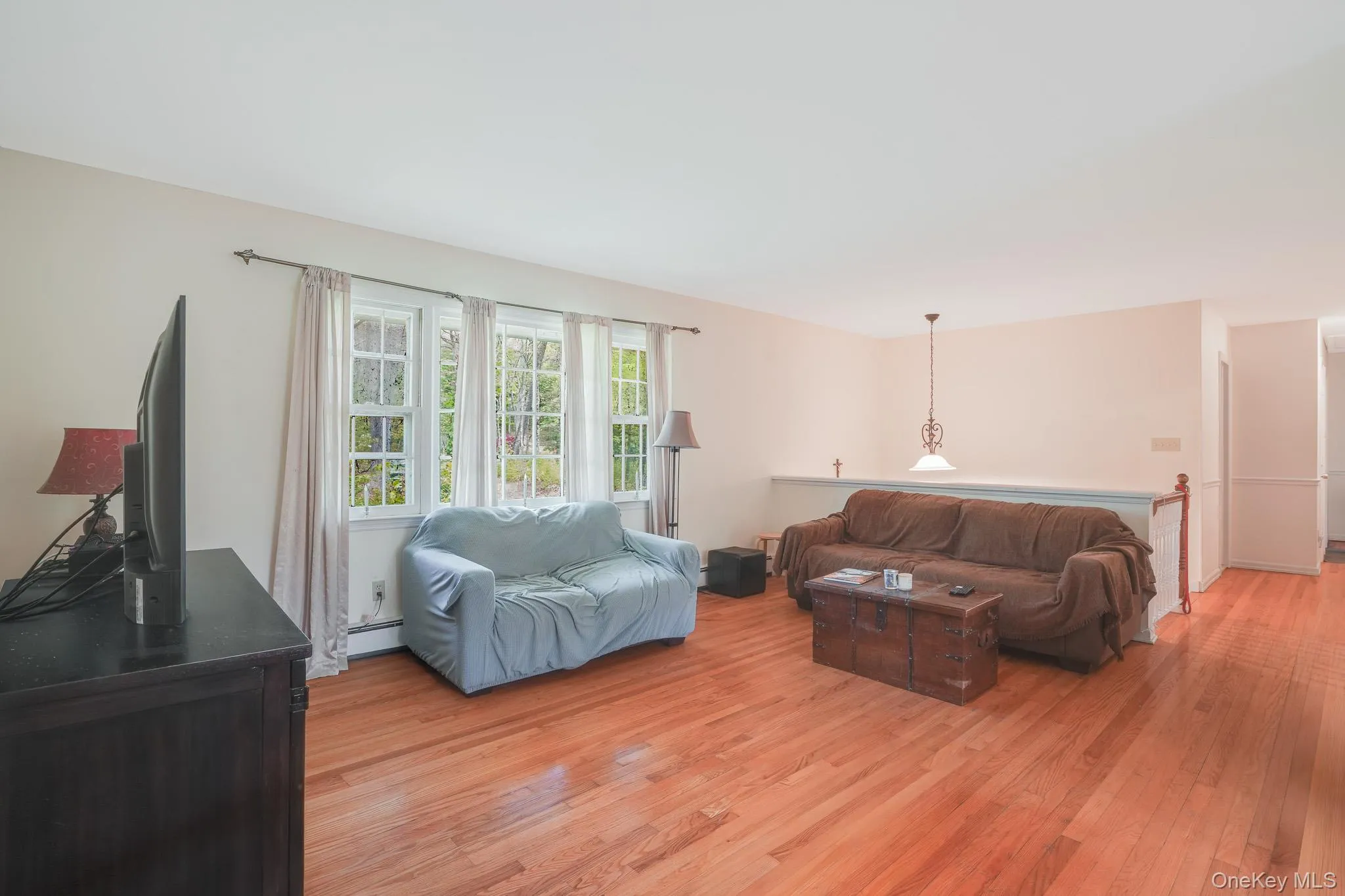 Hardwood flooring extends throughout this expansive living area, featuring a trio of windows, a half-wall partition, and a decorative pendant light fixture Hardwood flooring extends throughout this expansive living area, featuring a trio of windows, a half-wall partition, and a decorative pendant light fixture