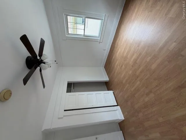 Bright room featuring wood-finish flooring, a ceiling fan with integrated lighting, and a window with white trim Bright room featuring wood-finish flooring, a ceiling fan with integrated lighting, and a window with white trim