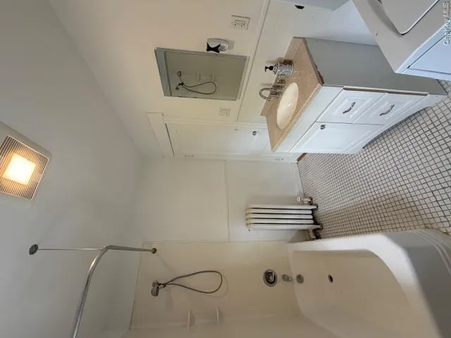 Bathroom featuring a white porcelain tub with shower, white vanity with integrated sink and stone-finish countertop, patterned floor tiling, and a wall-mounted radiator Bathroom featuring a white porcelain tub with shower, white vanity with integrated sink and stone-finish countertop, patterned floor tiling, and a wall-mounted radiator