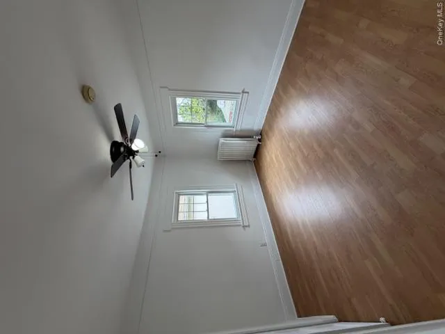 Room featuring wood-finish flooring, two windows with white trim, a ceiling fan with integrated lighting, and a white baseboard radiator Room featuring wood-finish flooring, two windows with white trim, a ceiling fan with integrated lighting, and a white baseboard radiator
