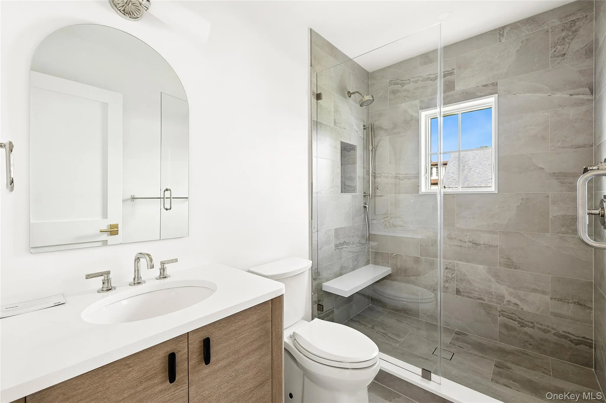 Contemporary bathroom featuring a floating wood-finish vanity with a white countertop and undermount sink, a large arch-top mirror, a frameless glass shower enclosure, a built-in shower niche, and a shower bench Contemporary bathroom featuring a floating wood-finish vanity with a white countertop and undermount sink, a large arch-top mirror, a frameless glass shower enclosure, a built-in shower niche, and a shower bench