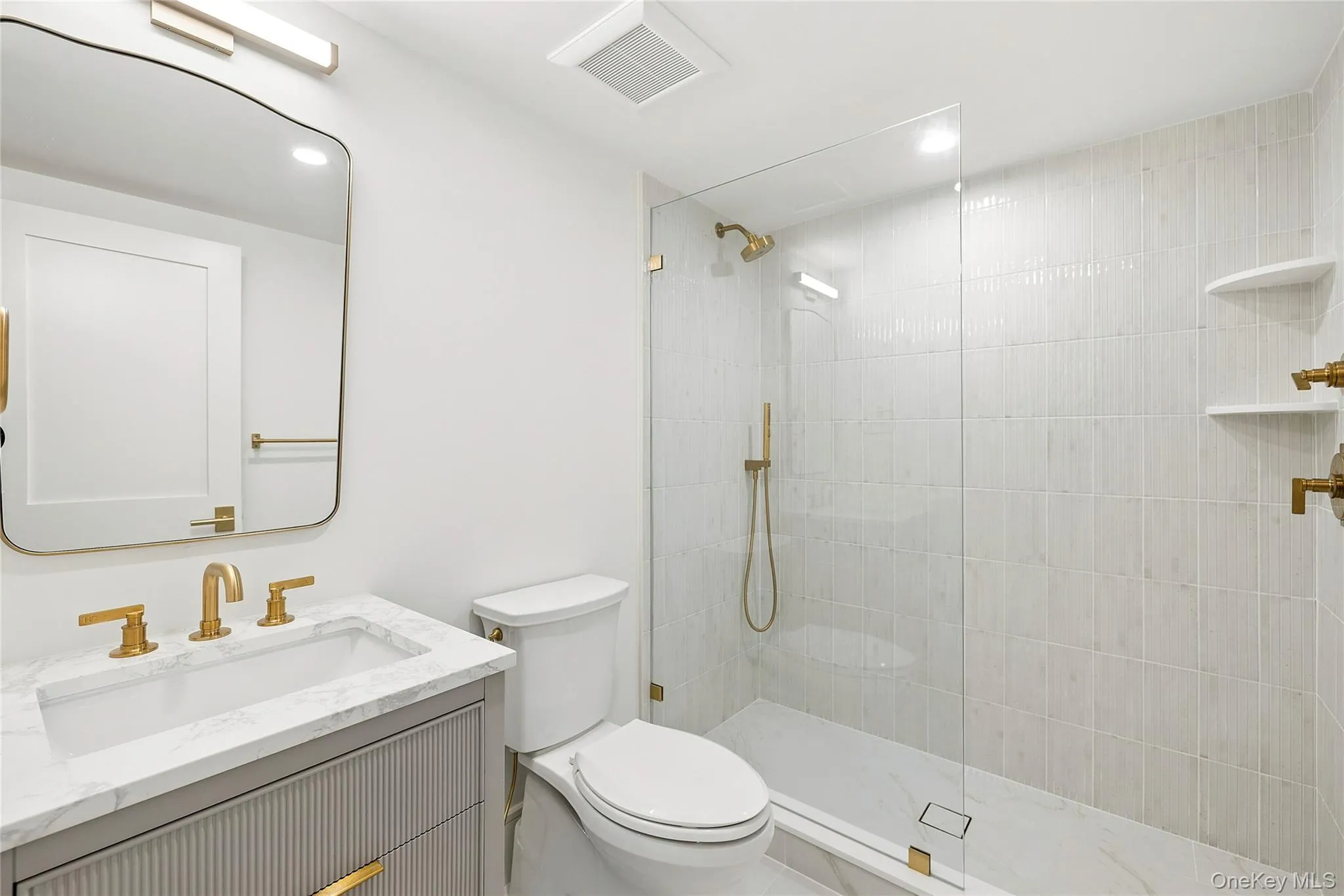 Contemporary bathroom featuring a glass-enclosed shower, gold-toned fixtures, a sleek vanity with a rectangular basin, a wall-mounted mirror, and white tiled walls Contemporary bathroom featuring a glass-enclosed shower, gold-toned fixtures, a sleek vanity with a rectangular basin, a wall-mounted mirror, and white tiled walls