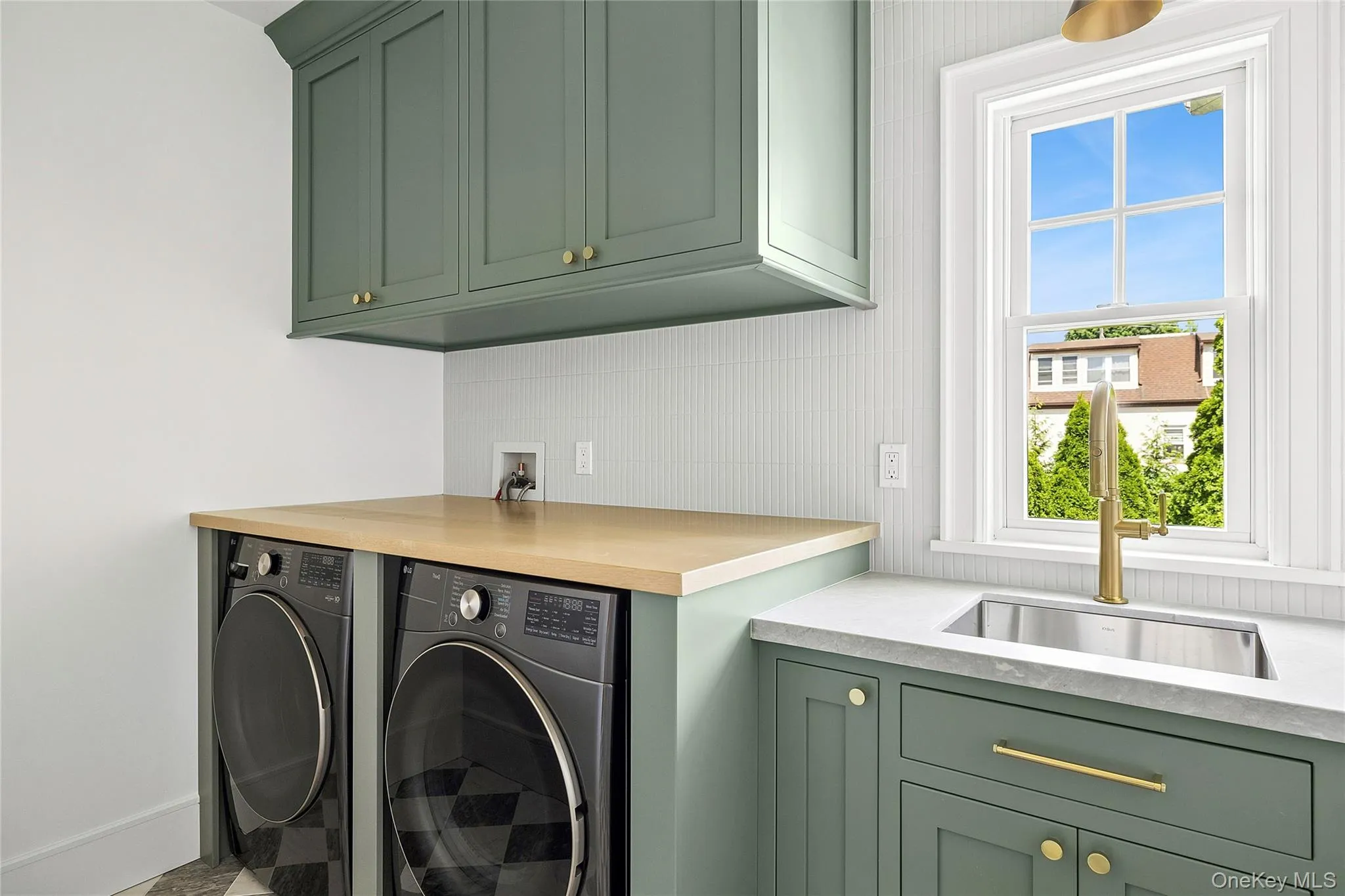 Dedicated laundry area featuring custom green cabinetry, a built-in sink with brass hardware, a light wood-finish countertop, and a white tiled backsplash Dedicated laundry area featuring custom green cabinetry, a built-in sink with brass hardware, a light wood-finish countertop, and a white tiled backsplash
