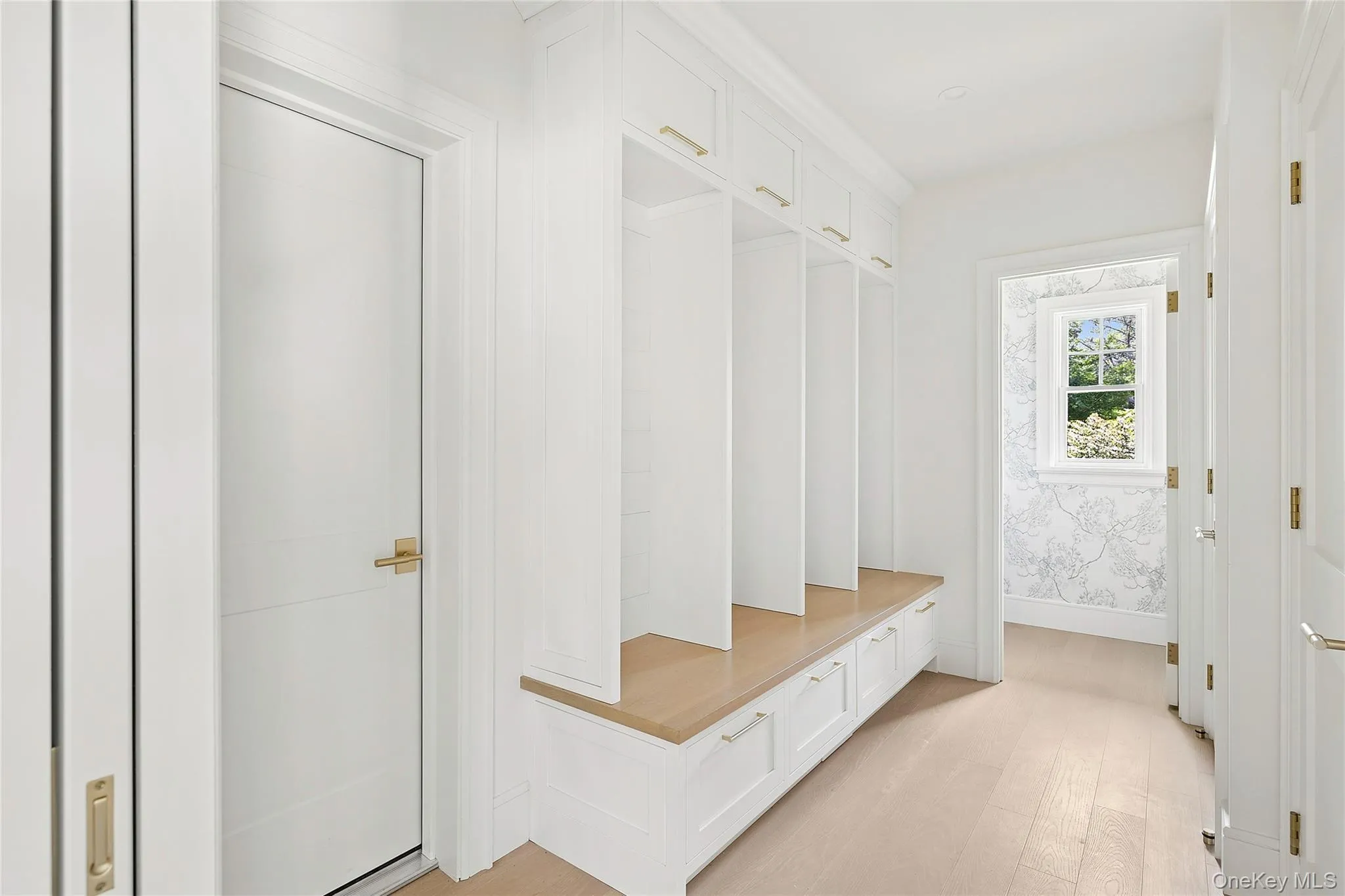 Mudroom featuring built-in white cabinetry with gold-tone hardware, a light wood-finish bench, and light wood-finish flooring Mudroom featuring built-in white cabinetry with gold-tone hardware, a light wood-finish bench, and light wood-finish flooring