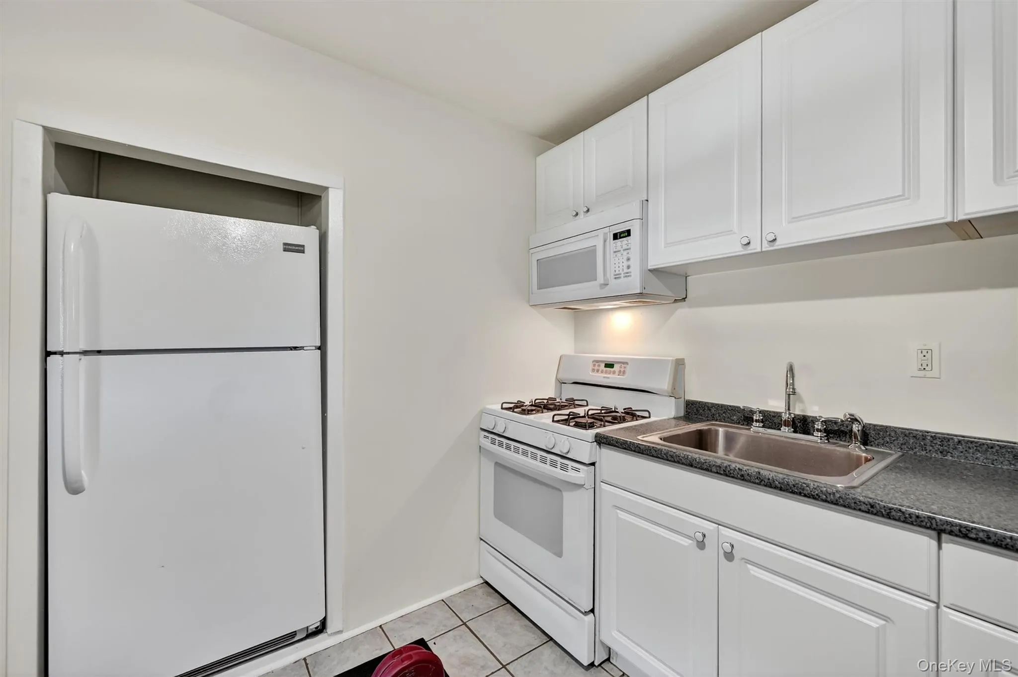 Kitchen featuring dark countertops, white appliances, white cabinets, and light tile patterned floors Kitchen featuring dark countertops, white appliances, white cabinets, and light tile patterned floors