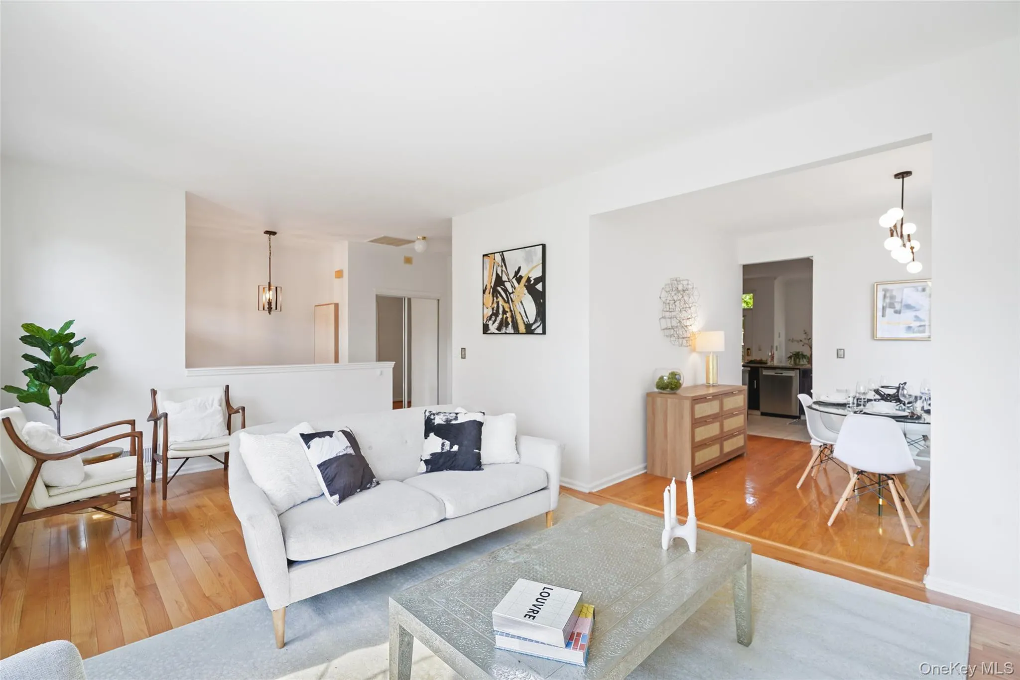 Bright living area featuring wood-finish flooring, white walls, and a recessed dining area with a modern chandelier Bright living area featuring wood-finish flooring, white walls, and a recessed dining area with a modern chandelier