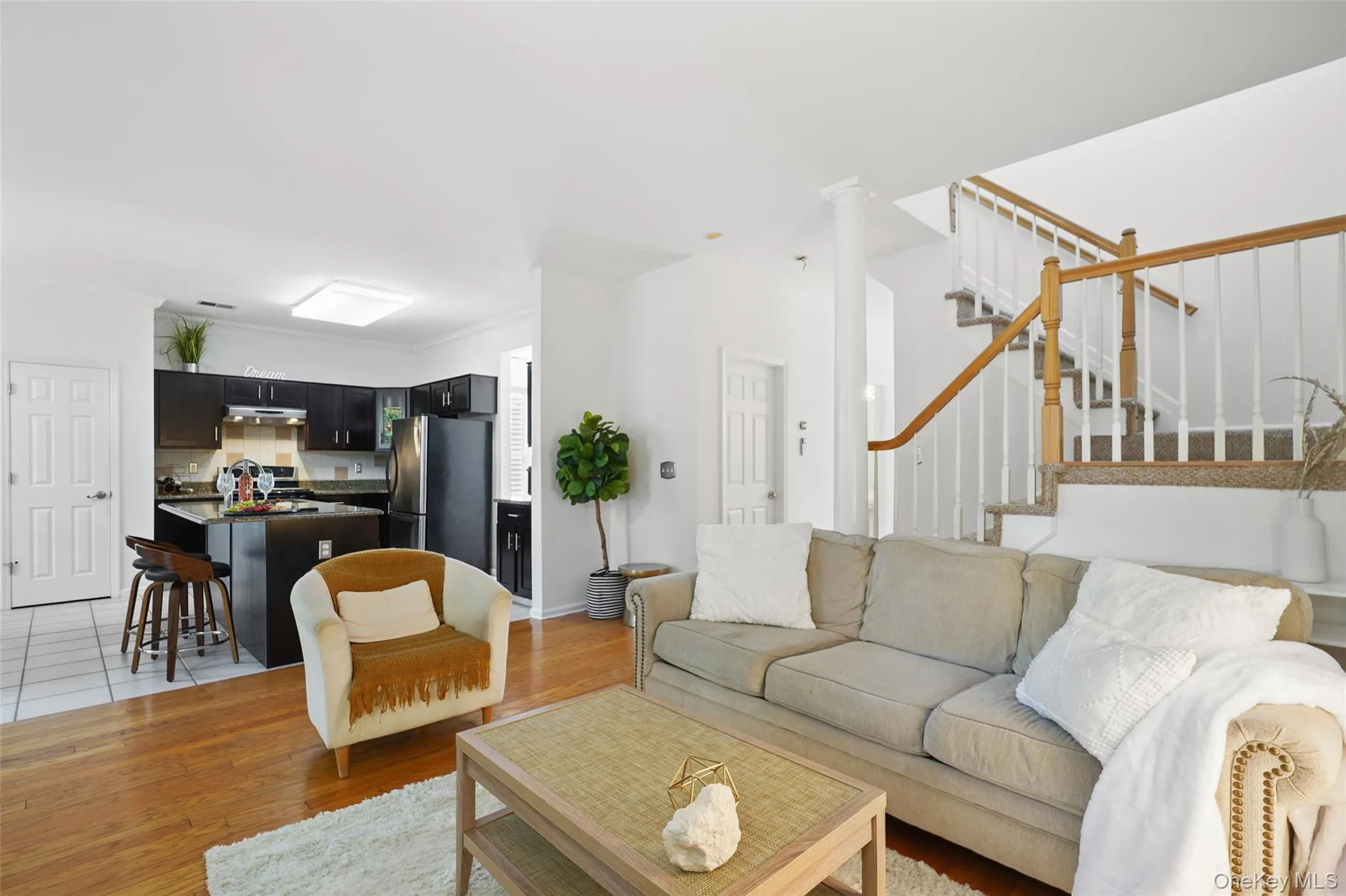 Open-concept living area featuring rich wood-finish flooring, a contemporary staircase with wood handrails and white balusters, and a kitchen with dark cabinetry, a central island, and stainless steel appliances Open-concept living area featuring rich wood-finish flooring, a contemporary staircase with wood handrails and white balusters, and a kitchen with dark cabinetry, a central island, and stainless steel appliances