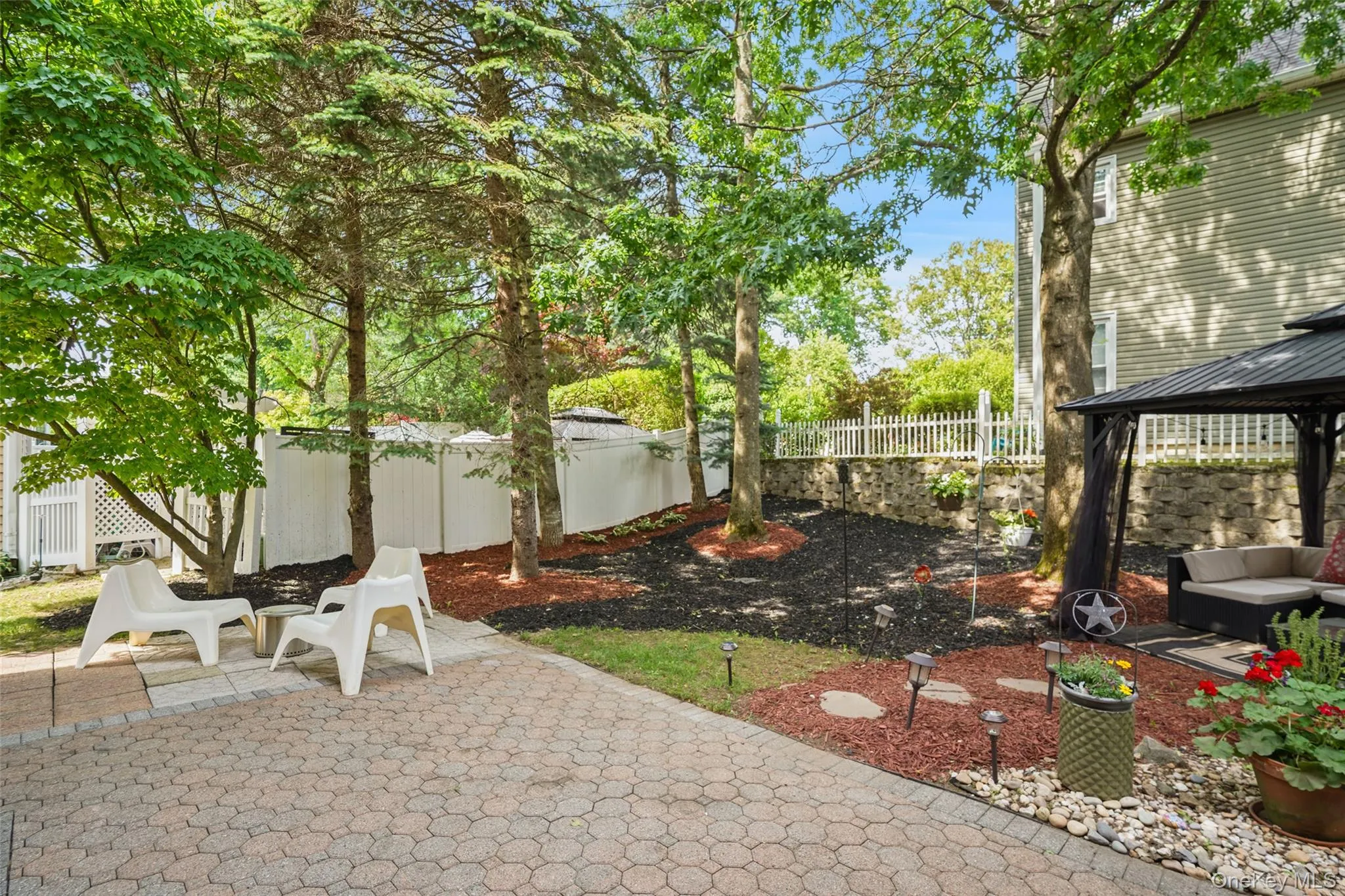 Paver patio providing an outdoor leisure space, surrounded by mature trees, a white privacy fence, and a stone retaining wall Paver patio providing an outdoor leisure space, surrounded by mature trees, a white privacy fence, and a stone retaining wall
