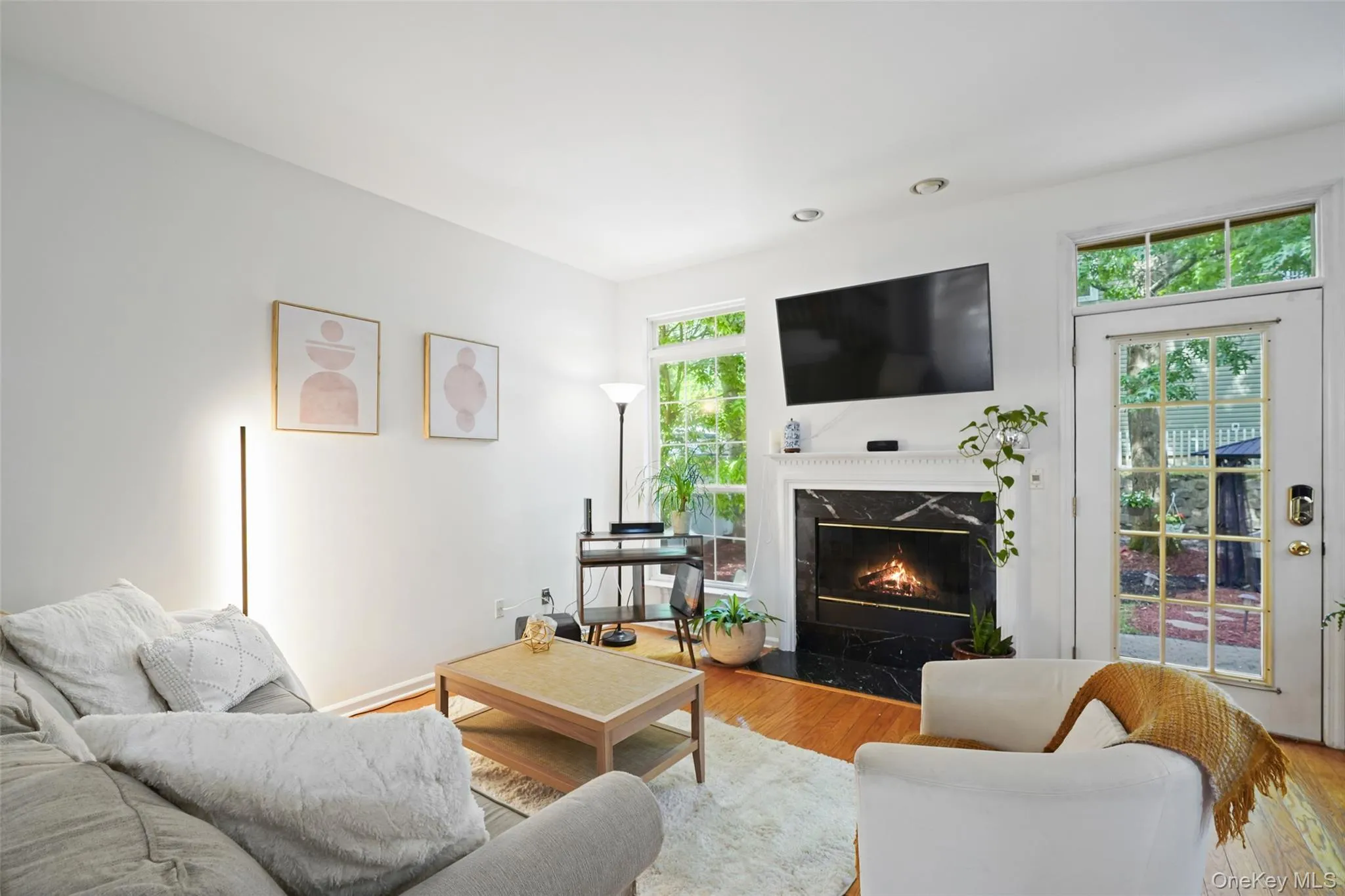 Living area featuring warm wood-finish flooring, a black marble-surround fireplace with a white mantel, and large windows providing natural light Living area featuring warm wood-finish flooring, a black marble-surround fireplace with a white mantel, and large windows providing natural light