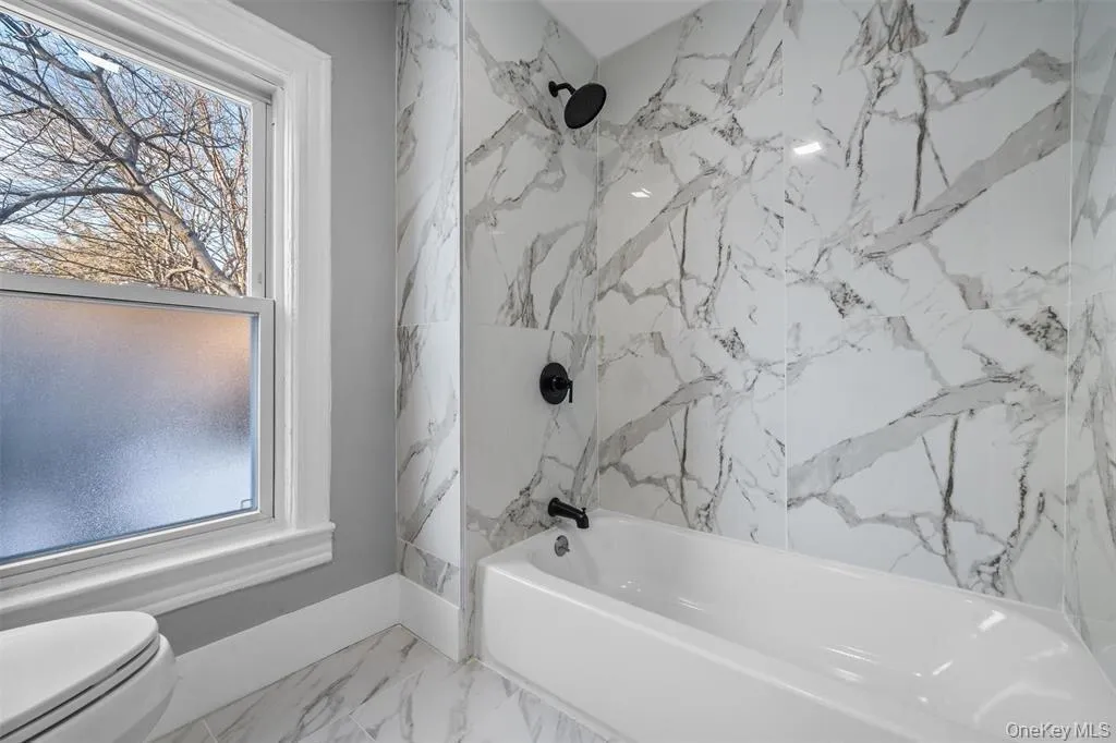 Bathroom featuring a white bathtub with matte black fixtures, floor-to-ceiling marbled tile, and a double-hung window with frosted glass Bathroom featuring a white bathtub with matte black fixtures, floor-to-ceiling marbled tile, and a double-hung window with frosted glass
