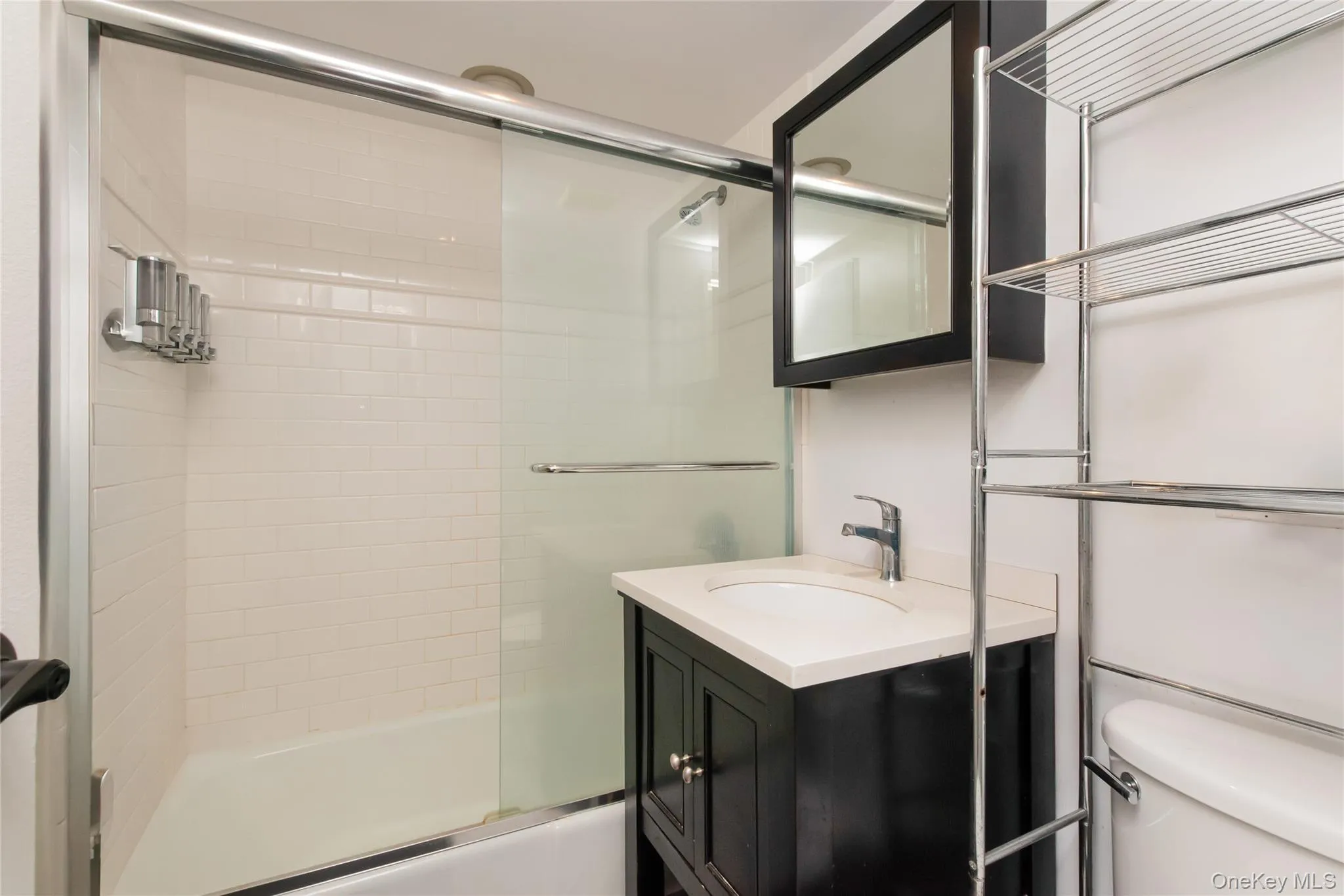 Bathroom featuring a white tile shower surround with a sliding glass door, a dark wood-finish vanity with a white countertop and undermount sink, and a mirrored medicine cabinet Bathroom featuring a white tile shower surround with a sliding glass door, a dark wood-finish vanity with a white countertop and undermount sink, and a mirrored medicine cabinet