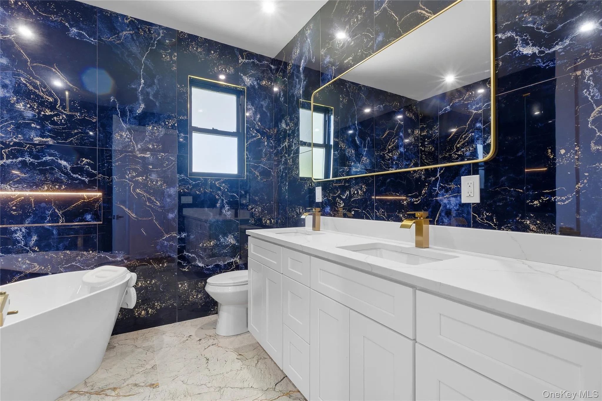 Bathroom featuring blue marble-effect wall tiles, a freestanding soaking tub, a double vanity with white cabinetry and integrated sinks, a gold-framed mirror, and gold-finish fixtures Bathroom featuring blue marble-effect wall tiles, a freestanding soaking tub, a double vanity with white cabinetry and integrated sinks, a gold-framed mirror, and gold-finish fixtures