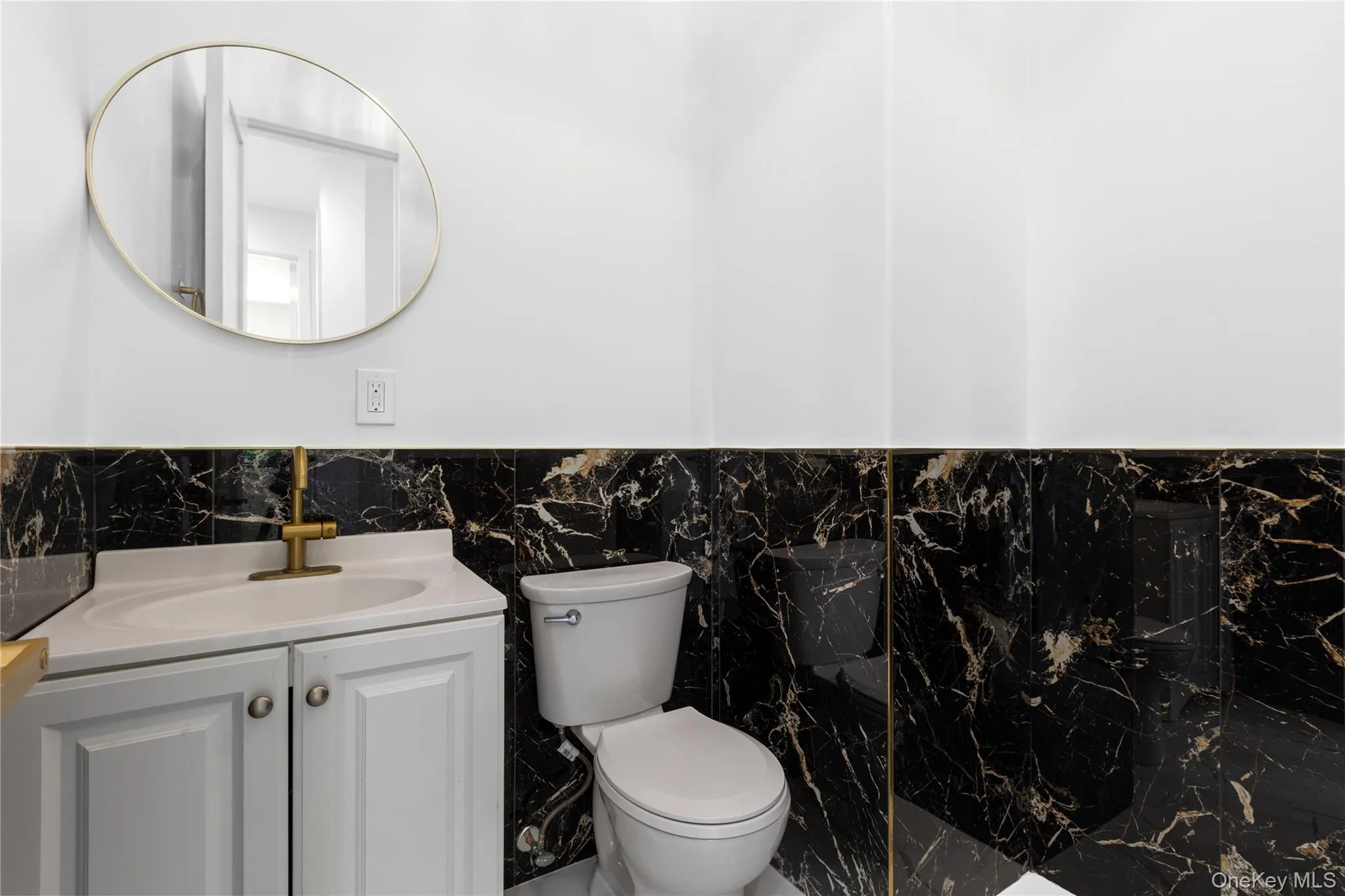 Powder room featuring black marble-style wall tiling, a white vanity with integrated sink, a gold-toned faucet, and a round wall mirror with a gold-tone frame Powder room featuring black marble-style wall tiling, a white vanity with integrated sink, a gold-toned faucet, and a round wall mirror with a gold-tone frame