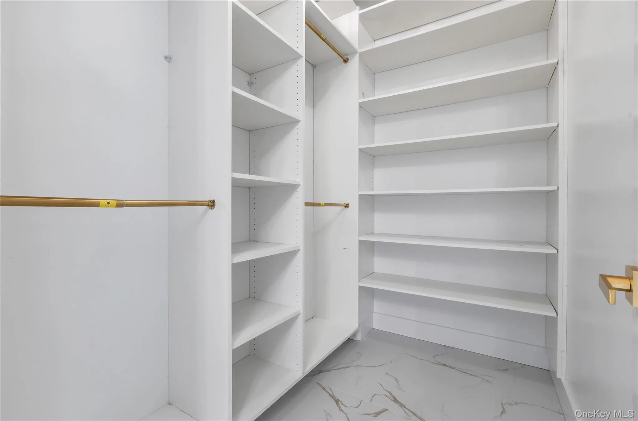 White custom closet system featuring multiple shelving units, brass-finish hanging rods, and light-toned flooring with subtle veining White custom closet system featuring multiple shelving units, brass-finish hanging rods, and light-toned flooring with subtle veining