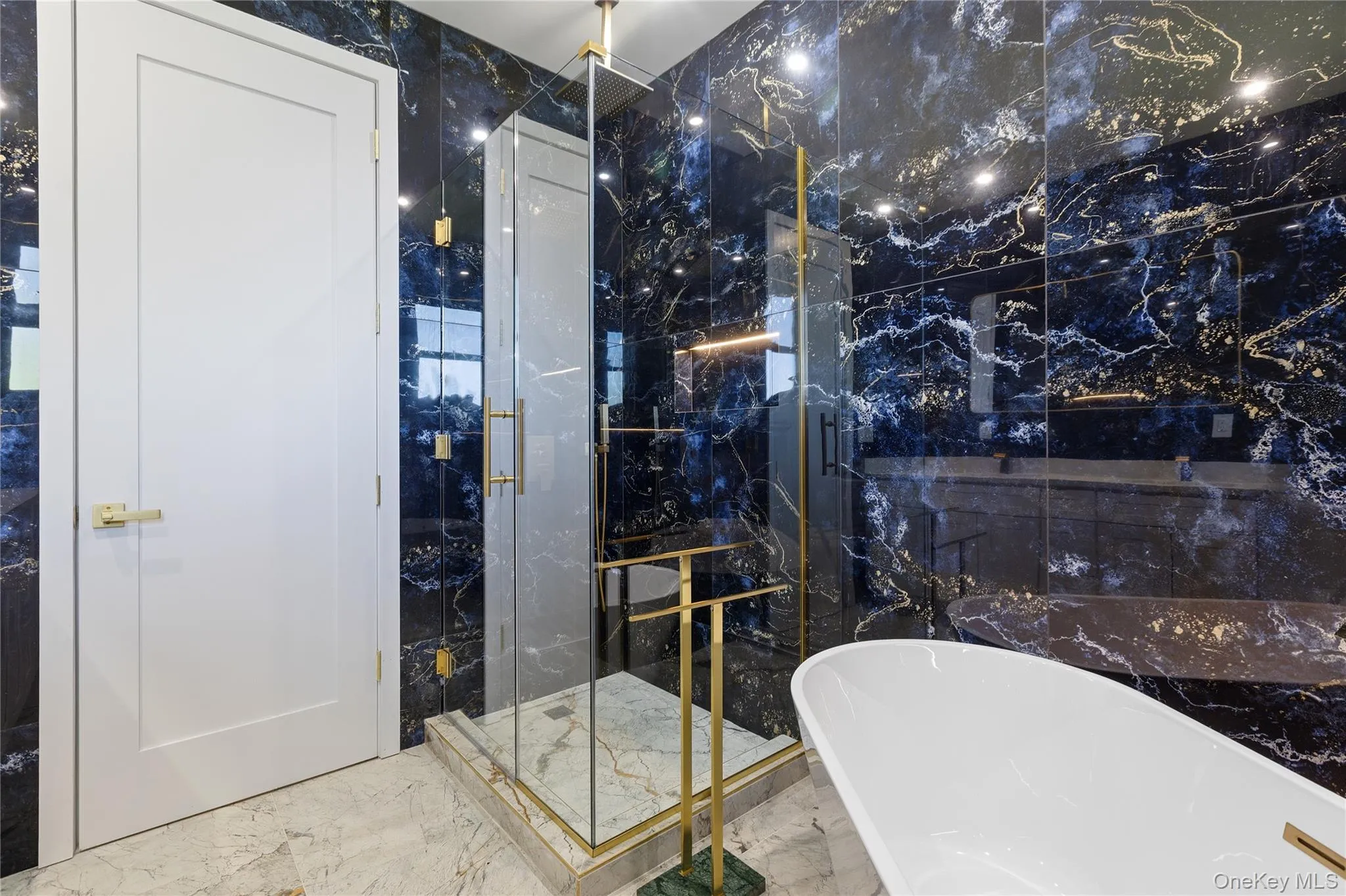 Bathroom featuring dark marbled wall tiles, a freestanding soaking tub, a glass-enclosed shower with brass-finish framing, and a white panel door with brass hardware Bathroom featuring dark marbled wall tiles, a freestanding soaking tub, a glass-enclosed shower with brass-finish framing, and a white panel door with brass hardware