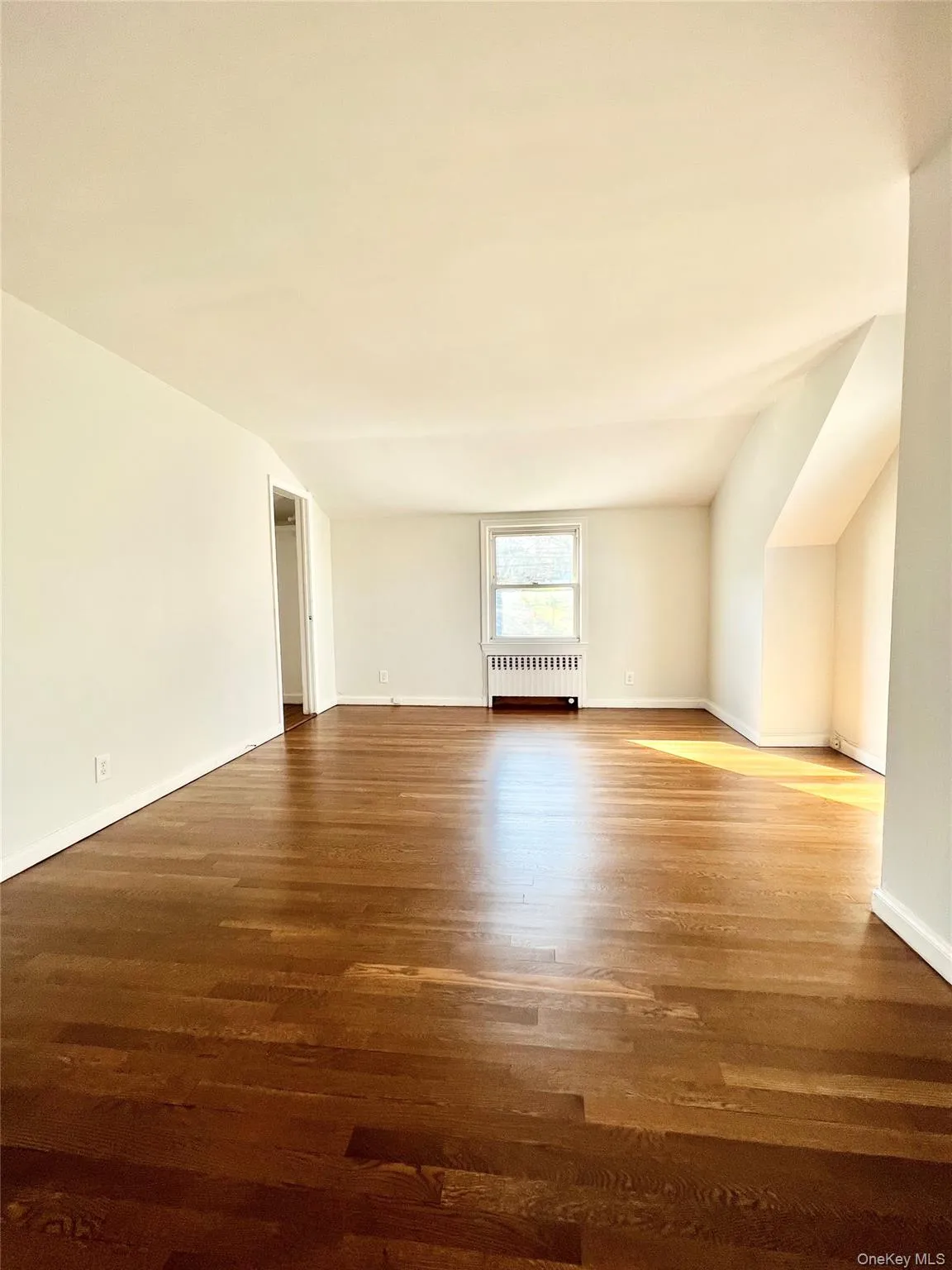 Spacious bedroom featuring rich wood-finish flooring, white walls, and a window providing natural illumination Spacious bedroom featuring rich wood-finish flooring, white walls, and a window providing natural illumination