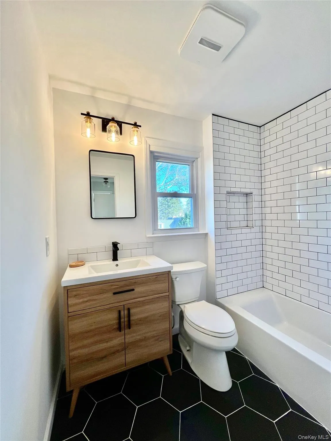 Bathroom featuring dark hexagonal floor tiles, a white subway-tiled shower with an integrated niche, and a light wood-finish vanity with a white countertop and rectangular sink Bathroom featuring dark hexagonal floor tiles, a white subway-tiled shower with an integrated niche, and a light wood-finish vanity with a white countertop and rectangular sink