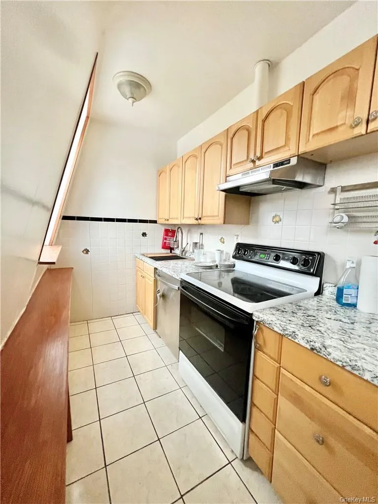 Kitchen featuring light wood-finish cabinetry, white tile flooring, a white tile backsplash with black accent trim, granite-patterned countertops, and a full-size electric range Kitchen featuring light wood-finish cabinetry, white tile flooring, a white tile backsplash with black accent trim, granite-patterned countertops, and a full-size electric range