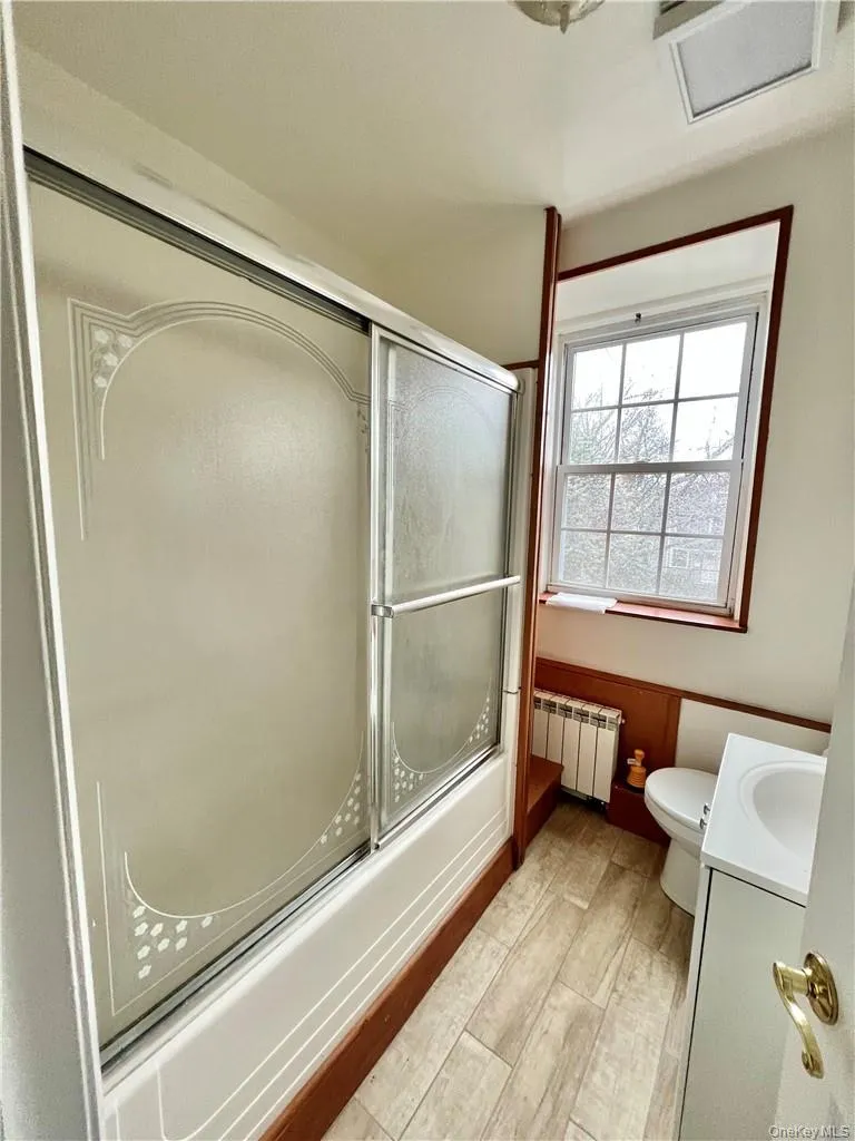 Bathroom featuring a tub/shower combination with sliding frosted glass doors, wood-finish flooring, a window providing natural light, a white vanity with an integrated sink, and a commode Bathroom featuring a tub/shower combination with sliding frosted glass doors, wood-finish flooring, a window providing natural light, a white vanity with an integrated sink, and a commode