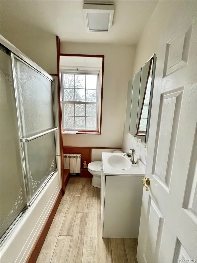 Compact bathroom featuring a white vanity with integrated sink, a mirrored medicine cabinet, a window with a wood frame, and wood-finish flooring Compact bathroom featuring a white vanity with integrated sink, a mirrored medicine cabinet, a window with a wood frame, and wood-finish flooring