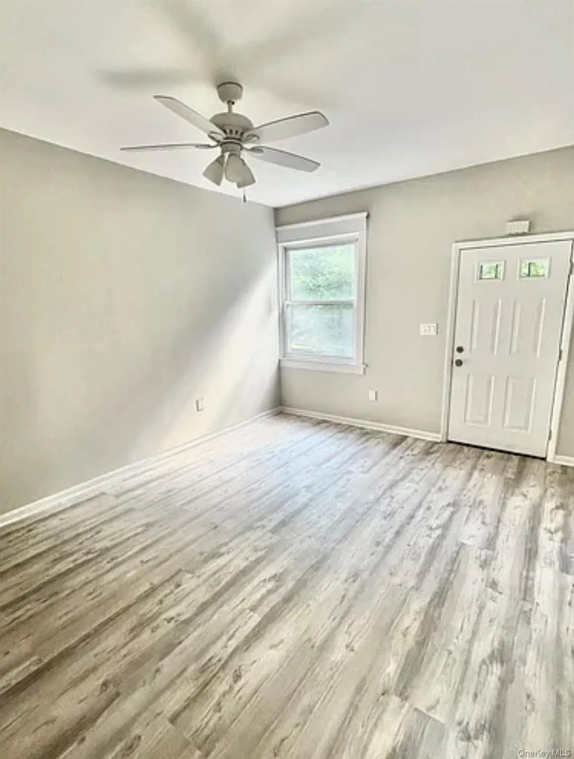 Spacious room featuring wood-finish flooring, light gray wall paint, a window providing ambient light, a ceiling fan with light fixture, and a white entry door with glass inserts Spacious room featuring wood-finish flooring, light gray wall paint, a window providing ambient light, a ceiling fan with light fixture, and a white entry door with glass inserts