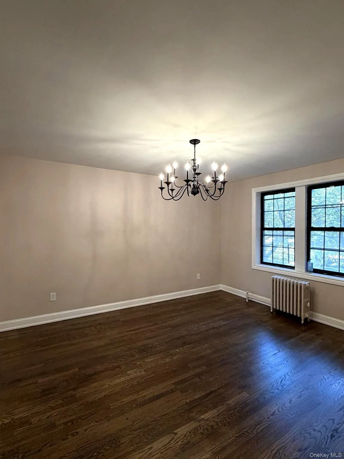 Spacious room featuring dark wood-finish flooring, a multi-arm chandelier, and two large windows with dark frames Spacious room featuring dark wood-finish flooring, a multi-arm chandelier, and two large windows with dark frames