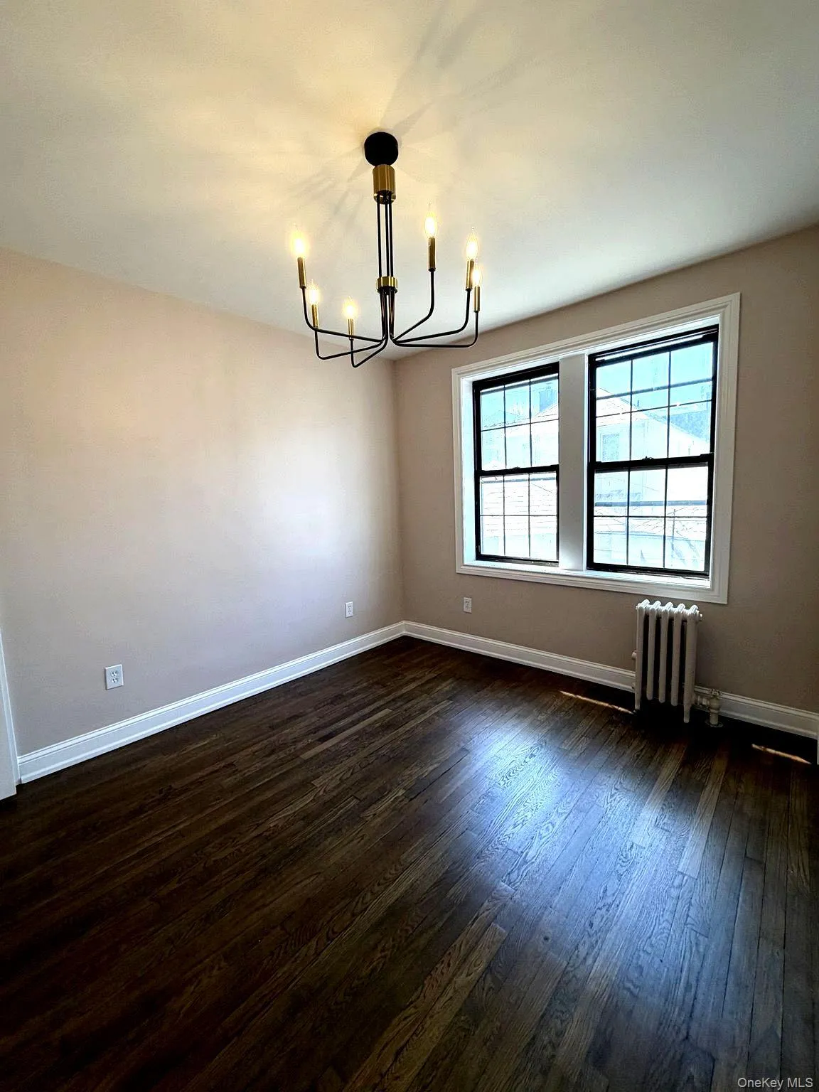 Room featuring dark wood-finish flooring, a contemporary six-light chandelier with black and brass accents, dual window units with black frames, and white baseboards Room featuring dark wood-finish flooring, a contemporary six-light chandelier with black and brass accents, dual window units with black frames, and white baseboards
