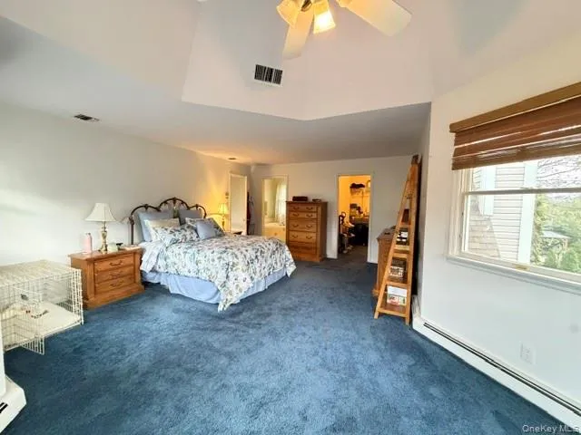 Spacious sleeping area featuring a vaulted ceiling with a ceiling fan, plush blue carpeting, a large window with privacy blinds, and baseboard heating Spacious sleeping area featuring a vaulted ceiling with a ceiling fan, plush blue carpeting, a large window with privacy blinds, and baseboard heating