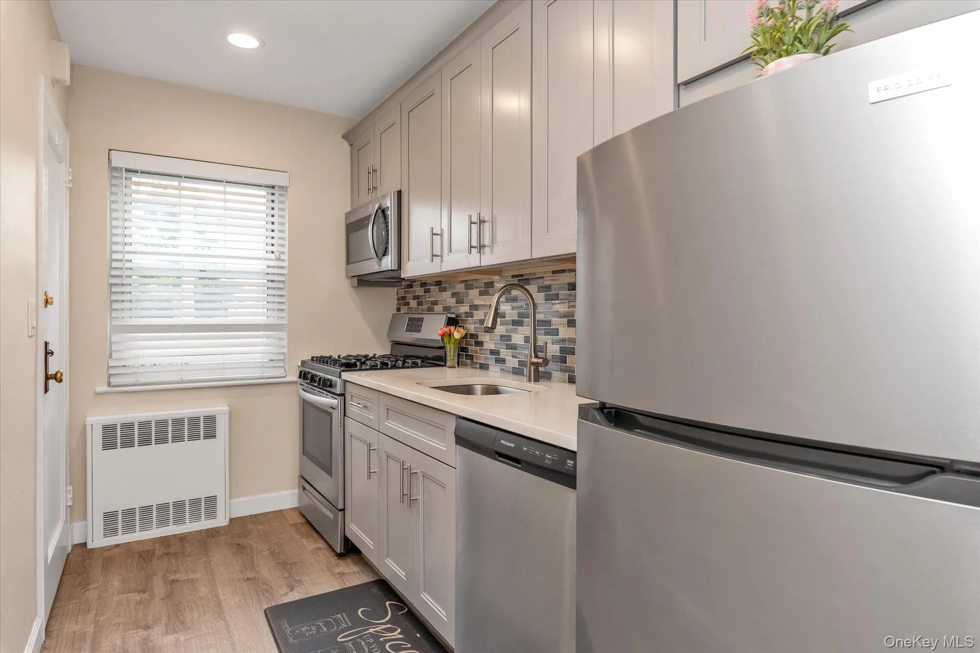 Kitchen featuring wood-finish flooring, stainless steel appliances, light-toned cabinetry, a mosaic tile backsplash, and a solid surface countertop Kitchen featuring wood-finish flooring, stainless steel appliances, light-toned cabinetry, a mosaic tile backsplash, and a solid surface countertop