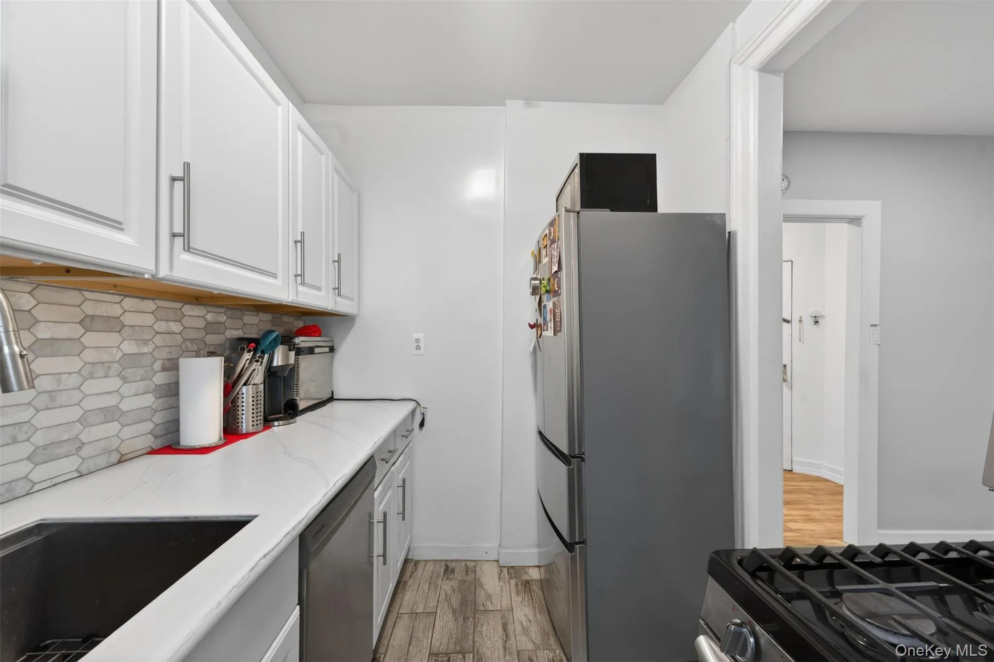 Galley-style kitchen featuring white upper cabinetry with brushed nickel hardware, a stainless steel sink, and wood-finish flooring Galley-style kitchen featuring white upper cabinetry with brushed nickel hardware, a stainless steel sink, and wood-finish flooring
