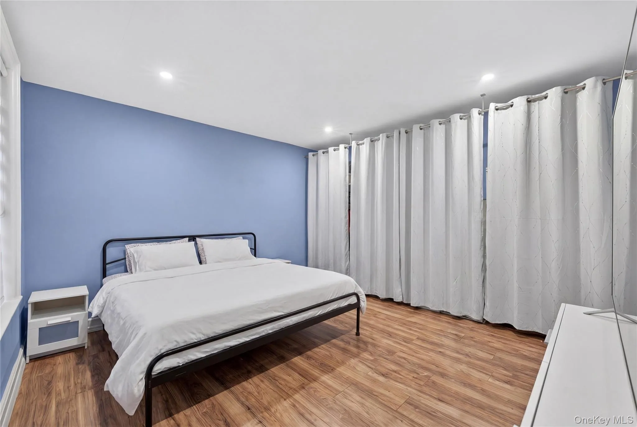 Room featuring wood-finish flooring, a light blue accent wall, recessed ceiling lighting, a white window frame, and a full wall of white privacy curtains Room featuring wood-finish flooring, a light blue accent wall, recessed ceiling lighting, a white window frame, and a full wall of white privacy curtains