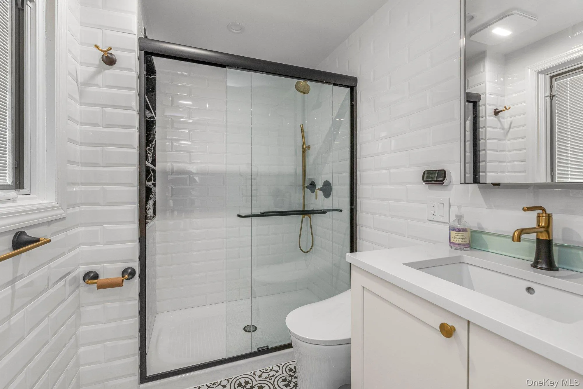 Bathroom featuring white subway tile walls, a frameless glass shower enclosure with black trim, a vanity with a white countertop and integrated sink, a gold and black faucet, and patterned floor tiling Bathroom featuring white subway tile walls, a frameless glass shower enclosure with black trim, a vanity with a white countertop and integrated sink, a gold and black faucet, and patterned floor tiling