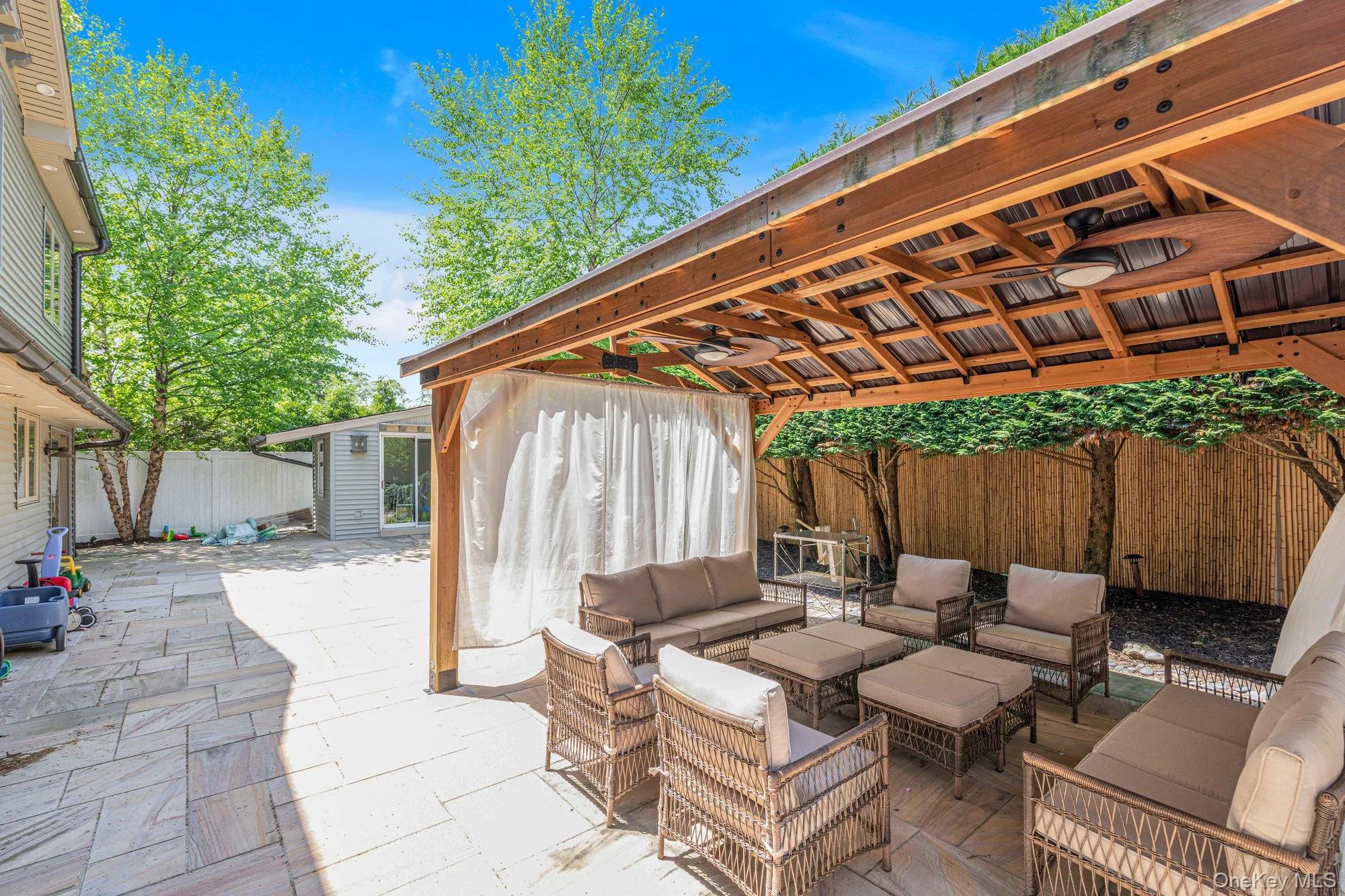 Expansive outdoor patio featuring a wood pergola with integrated ceiling fans, privacy curtains, and a stone paver surface Expansive outdoor patio featuring a wood pergola with integrated ceiling fans, privacy curtains, and a stone paver surface