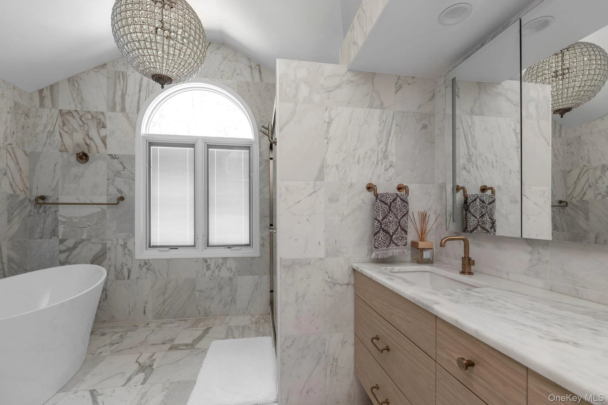Marble tiled bathroom featuring a freestanding soaking tub, an arched window, a crystal chandelier, a vanity with a light wood finish, and a coordinating marble countertop Marble tiled bathroom featuring a freestanding soaking tub, an arched window, a crystal chandelier, a vanity with a light wood finish, and a coordinating marble countertop