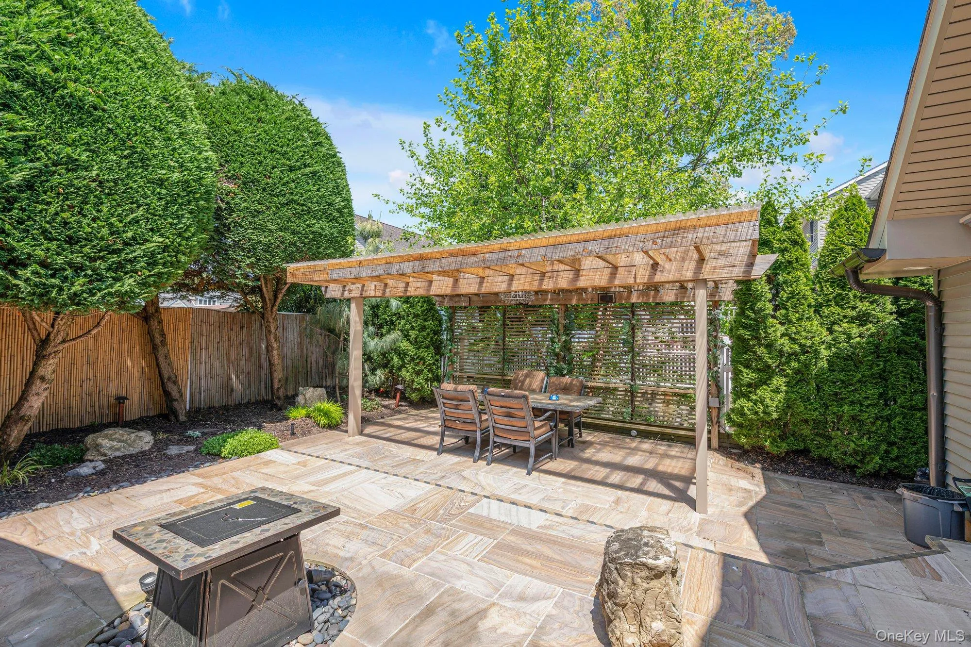 Outdoor patio featuring a wooden pergola, stone-finish paver flooring, a built-in fire pit, privacy fencing, and established perimeter plantings Outdoor patio featuring a wooden pergola, stone-finish paver flooring, a built-in fire pit, privacy fencing, and established perimeter plantings