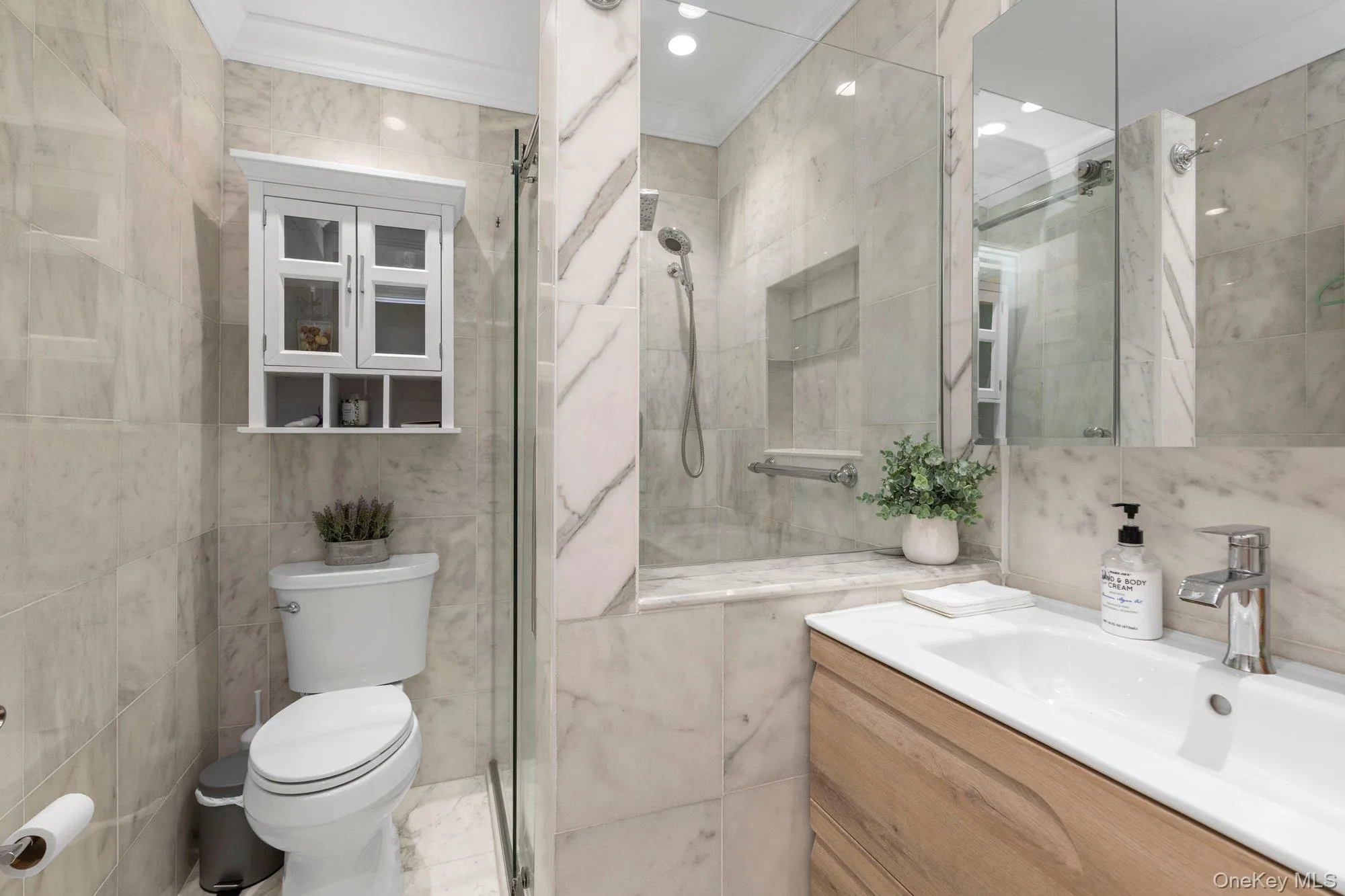 Bathroom featuring a walk-in shower with a glass enclosure, white rectangular under-mount sink, wood-finish vanity, wall-mounted mirrored medicine cabinet, and tiled walls and flooring Bathroom featuring a walk-in shower with a glass enclosure, white rectangular under-mount sink, wood-finish vanity, wall-mounted mirrored medicine cabinet, and tiled walls and flooring