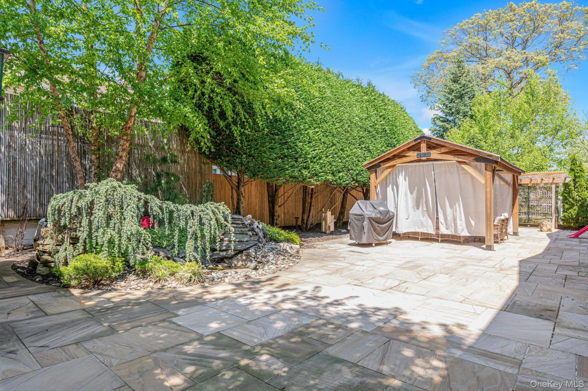 Expansive stone patio featuring a wooden gazebo with privacy drapes, dense green hedges, mature trees, and a decorative rock garden with abundant foliage Expansive stone patio featuring a wooden gazebo with privacy drapes, dense green hedges, mature trees, and a decorative rock garden with abundant foliage