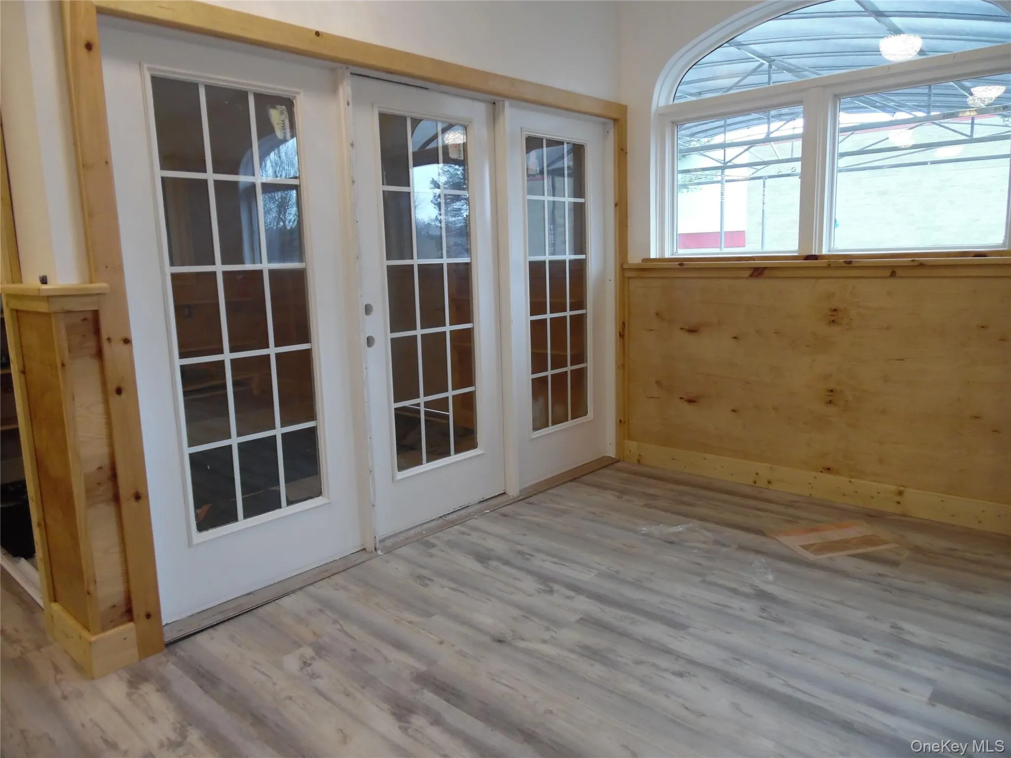 French doors with divided lites open onto a room featuring wood-finish flooring, a wood wainscoting detail, and a large arched window French doors with divided lites open onto a room featuring wood-finish flooring, a wood wainscoting detail, and a large arched window