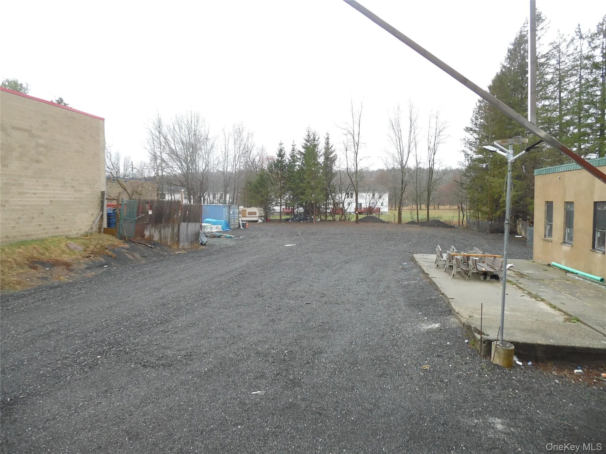 Gravel parking area featuring a concrete pad, surrounded by mature trees and a brick building Gravel parking area featuring a concrete pad, surrounded by mature trees and a brick building