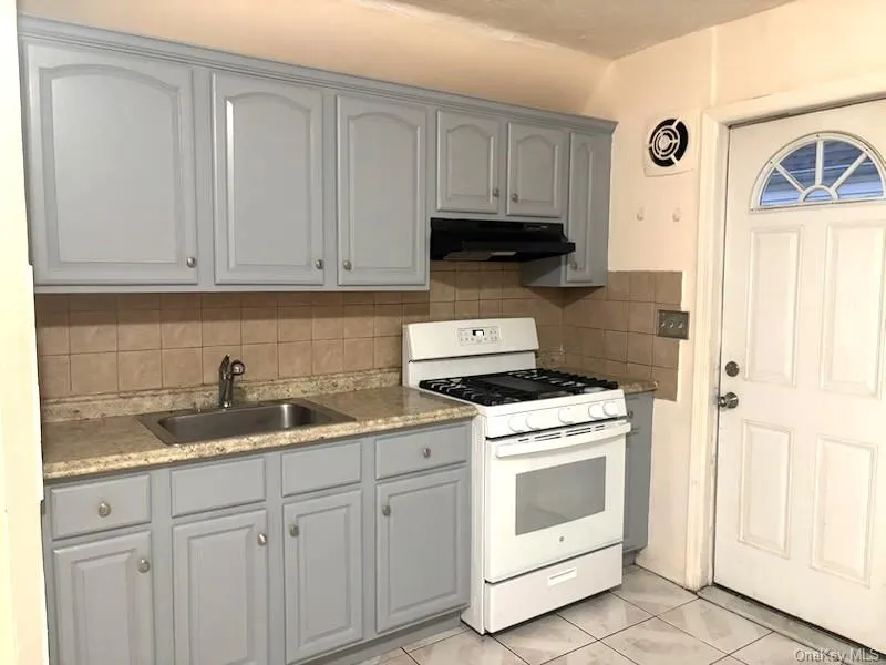 Kitchen featuring gray cabinets, white range with gas stovetop, tasteful backsplash, light tile patterned floors, and under cabinet range hood Kitchen featuring gray cabinets, white range with gas stovetop, tasteful backsplash, light tile patterned floors, and under cabinet range hood