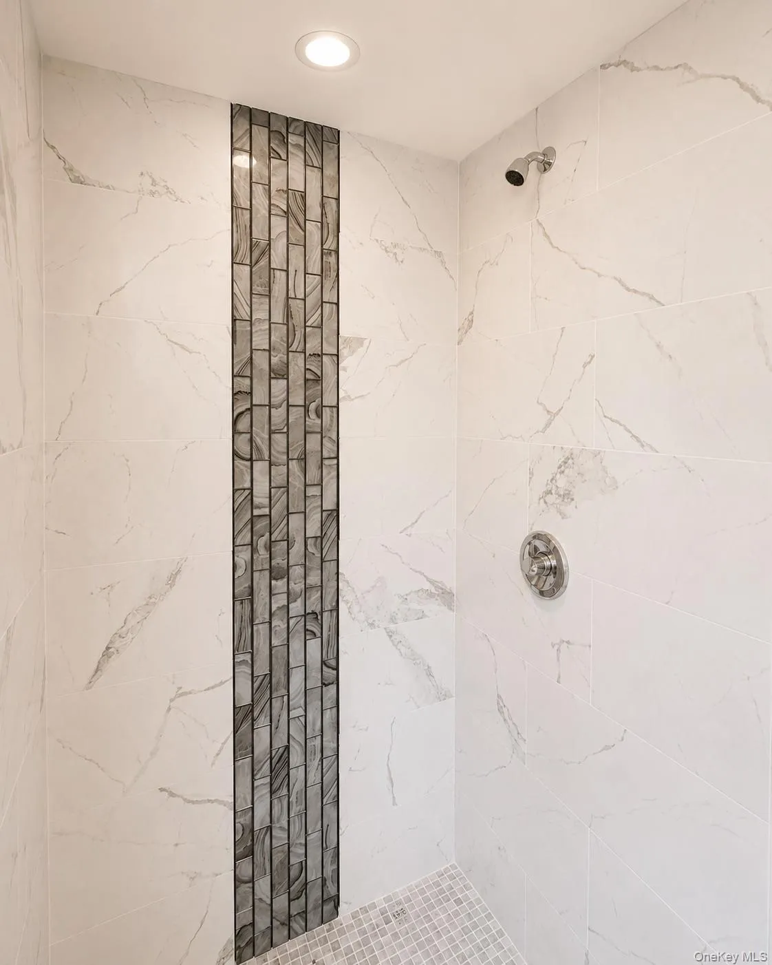 Shower enclosure featuring large format white tile walls with gray veining, a recessed ceiling light, and a vertical mosaic accent strip Shower enclosure featuring large format white tile walls with gray veining, a recessed ceiling light, and a vertical mosaic accent strip
