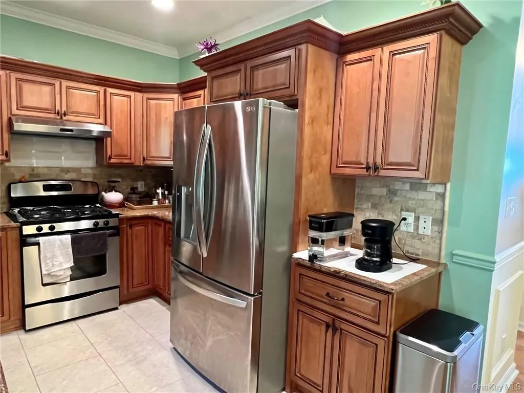 Kitchen with stainless steel appliances, under cabinet range hood, brown cabinetry, tasteful backsplash, and ornamental molding Kitchen with stainless steel appliances, under cabinet range hood, brown cabinetry, tasteful backsplash, and ornamental molding