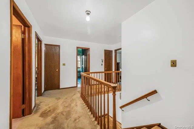 Upper-level hallway featuring a wooden banister, carpeted flooring, white walls, and multiple brown wood-paneled doors Upper-level hallway featuring a wooden banister, carpeted flooring, white walls, and multiple brown wood-paneled doors